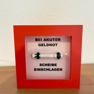May include: A red framed display box with the text "BEI AKUTER GELDNOT SCHEIBE EINSCHLAGEN" in black. Inside the box is a white object with black and green accents.