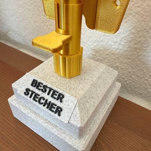 May include: A gold-colored trophy with a white base. The base has the words "BESTER STECHER" printed on it.