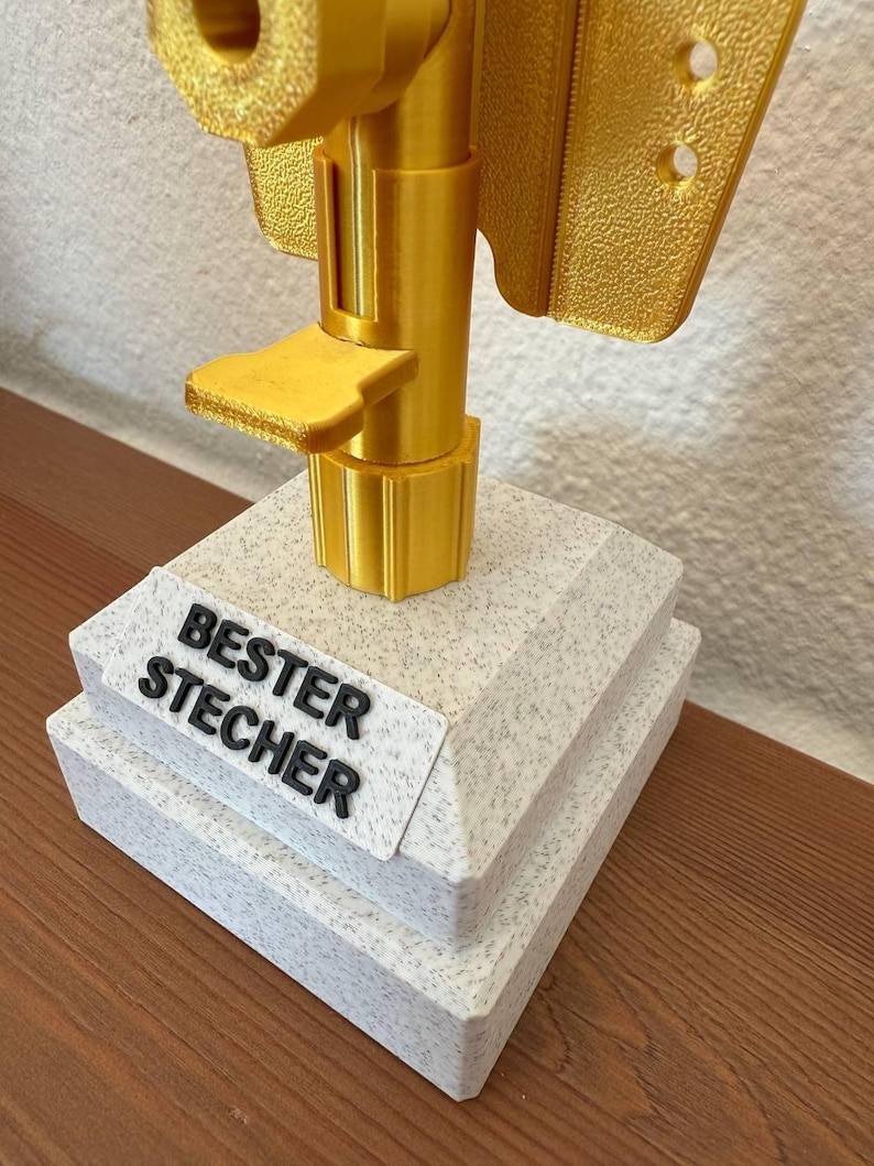 May include: A gold-colored trophy with a white base. The base has the words "BESTER STECHER" printed on it.