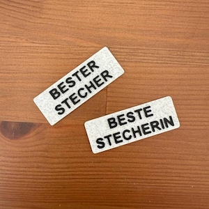 May include: Two white rectangular patches with black text. The top patch reads "BESTER STECHER" and the bottom patch reads "BESTE STECHERIN".