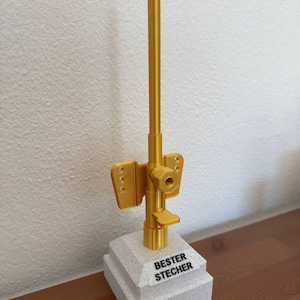 Könnte beinhalten: Eine goldfarbene Trophäe mit einem hohen, dünnen, zylindrischen Sockel und einem kleineren, quadratischen Sockel. Die Spitze der Trophäe ist ein langes, dünnes, goldfarbenes Rohr. Der quadratische Sockel trägt die Aufschrift "BESTER STECHER".