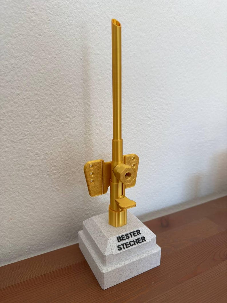 May include: A gold-colored trophy with a tall, thin, cylindrical base and a smaller, square base. The top of the trophy is a long, thin, gold-colored tube. The square base has the words "BESTER STECHER" printed on it.