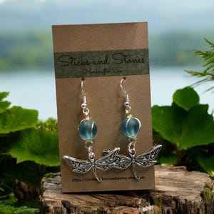 May include: A pair of silver dragonfly earrings with blue glass beads. The earrings are on a brown card with the text "Sticks and Stones Handmade Zen" and a website address.
