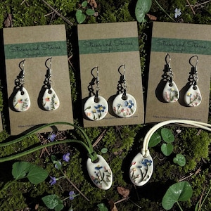May include: Handmade ceramic earrings and pendants featuring floral designs. The earrings are displayed on brown cards with the text "Sticks and Stones." The jewelry includes white, teardrop, and circular shapes with colorful flower details.