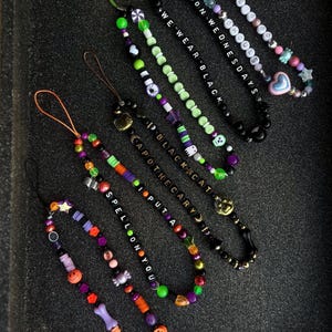 May include: Six colorful phone charms with black letter beads spelling out phrases like "Spell on you", "I Put a Spell on You", "Black Cat", "We Wear Black", and "On Wednesdays We Wear Black".