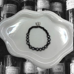 May include: Black beaded bracelet with a silver book charm. The beads are black and have white letters spelling out "Turn to Page 394".