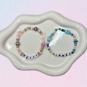 May include: Two beaded bracelets with the names "BETTY" and "JAMES" spelled out in white letters. The bracelets are on a white ceramic plate with a wavy edge.