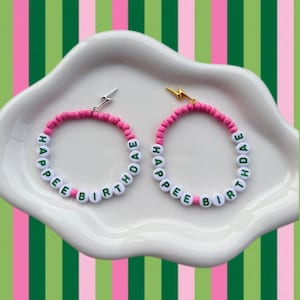 May include: Two bracelets with the text "HAPPY BIRTHDAE" in white letter beads. The bracelets have pink beads and silver and gold lightning bolt charms. The bracelets are on a white, cloud-shaped dish against a green, pink, and white striped background.