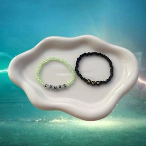 May include: Two beaded bracelets, one light green and one black, sit on a white, wavy dish. The light green bracelet spells "LUMOS" and the black bracelet spells "NOX". The bracelets are likely themed after a popular fantasy series.