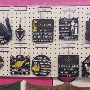May include: A white pegboard display featuring numerous black decorative wall plaques and ornaments with various yellow and white text and graphic designs, including quotes, humorous sayings, and symbols like horseshoes and hearts, all hanging on metal hooks.