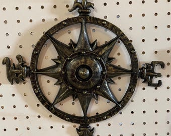 May include: A decorative metal compass rose with a dark bronze finish. The compass features directional indicators (N, E, S, W) and ornate detailing. It is designed for wall mounting and adds a vintage nautical touch to decor.