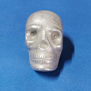 May include: A silver-colored skull figurine with detailed facial features, including eye sockets, a nose cavity, and a jawline. The skull is set against a blue background, creating a striking contrast. This decorative piece is likely used for Halloween or gothic-themed decor.