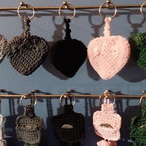 May include: A display of handmade crochet keychains in various colors, including gray, black, pink, and green. The keychains are heart-shaped and rectangular, each attached to a metal ring. They are arranged on a gold-colored metal rack against a blue background.