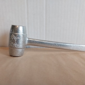 May include: A silver-colored nutcracker with a mallet-shaped head and a long handle. The head features an embossed floral design. The nutcracker is made of metal and is designed for cracking nuts.