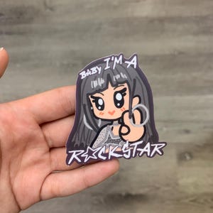 Op de afbeelding: Een sticker met een cartoon illustratie van een meisje met donker haar en grote ogen. De sticker heeft de tekst "BABY I'M A ROCKSTAR" in een gestileerd lettertype. Het meisje maakt een handgebaar.