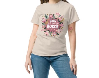 Könnte beinhalten: Beiges T-Shirt mit floralem Design und dem Text "Born to Bokkie" in Rosa und Weiß. Das Design beinhaltet rosa Blumen und grüne Blätter. Das Model trägt eine blaue Jeans.