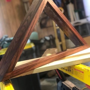 Könnte beinhalten: Ein dreieckiger Display-Rahmen aus dunklem Holz. Der Rahmen hat abgewinkelte Seiten, die ein Dreieck bilden. Das Gehäuse ruht auf einer hellen Holzoberfläche. Das Holz hat eine polierte Oberfläche.