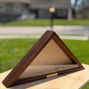Puede incluir: Una vitrina triangular de madera con marco marrón oscuro e interior claro. Una pequeña placa rectangular dorada es visible en el borde inferior. Diseñada para exhibir una bandera.