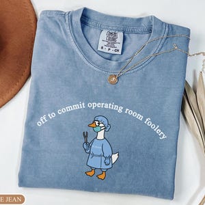 May include: A blue jean colored t-shirt featuring a graphic of a goose dressed in surgical scrubs, a mask, and glasses, holding a surgical tool. The text above the goose reads, "off to commit operating room foolery." The shirt has a Comfort Colors label.