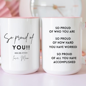 May include: Two white ceramic mugs with pink handles and rims. One mug says "So proud of YOU!!" with "love, Mom" and a date. The other mug says "So proud of who you are..." and "of all you have accomplished."