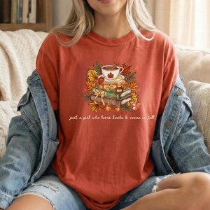 May include: A rust-colored t-shirt featuring a graphic of a stack of books topped with a cup of cocoa, surrounded by autumn leaves and a small squirrel. The text reads, "just a girl who loves books & cocoa in fall" in a white cursive script.