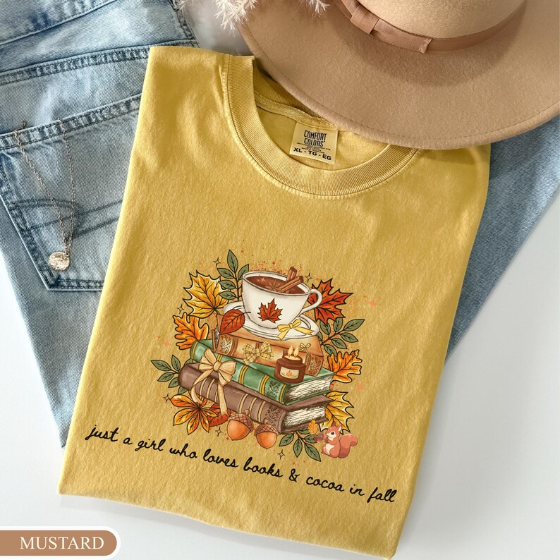 May include: A mustard yellow t-shirt featuring a graphic of a stack of books, a cup of coffee, autumn leaves, and a squirrel. The text reads, "just a girl who loves books & cocoa in fall." The shirt has a crew neckline and a Comfort Colors brand label.