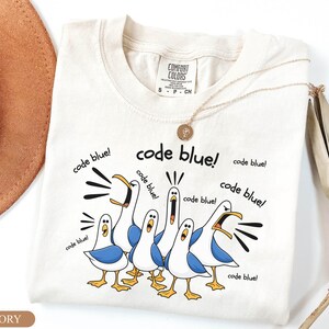 May include: An ivory short-sleeve t-shirt featuring a graphic of five white and blue seagulls with yellow feet and beaks. The seagulls are depicted with open mouths and the text "code blue!" is printed multiple times around the birds in black.