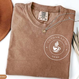 May include: A brown short-sleeved t-shirt with a crew neckline and a Comfort Colors label. The shirt features a white circular graphic on the chest containing a coffee cup icon and the text CUTOSM LOGO HERE. A silver chain necklace rests on top.