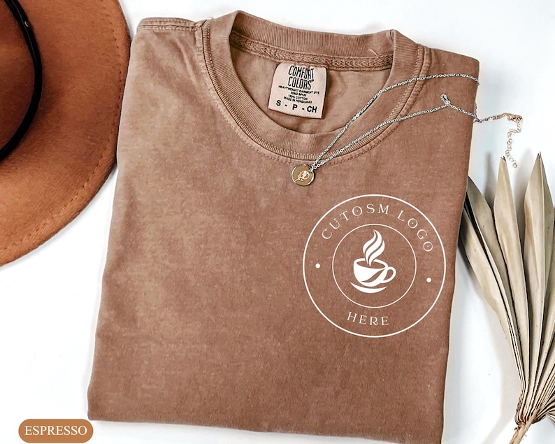 May include: A brown short-sleeved t-shirt with a crew neckline and a Comfort Colors label. The shirt features a white circular graphic on the chest containing a coffee cup icon and the text CUTOSM LOGO HERE. A silver chain necklace rests on top.