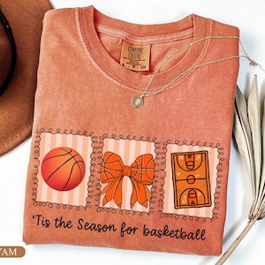 Peut inclure: Un t-shirt corail avec un motif sur le th&egrave;me du basketball. Le motif comprend un ballon de basket, un n&oelig;ud et un terrain de basket, avec le texte "'Tis the Season for basketball". Un collier en or est &eacute;galement visible.
