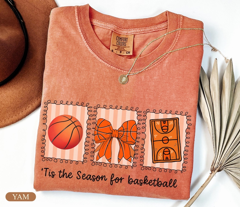 Peut inclure: Un t-shirt corail avec un motif sur le th&egrave;me du basketball. Le motif comprend un ballon de basket, un n&oelig;ud et un terrain de basket, avec le texte "'Tis the Season for basketball". Un collier en or est &eacute;galement visible.
