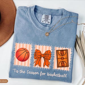 Peut inclure: T-shirt bleu jean clair avec des graphiques sur le th&egrave;me du basket-ball : un ballon, un n&oelig;ud et un terrain. Le texte "'Tis the Season for basketball" est imprim&eacute; en dessous. Un collier dor&eacute; est drap&eacute; sur le t-shirt.