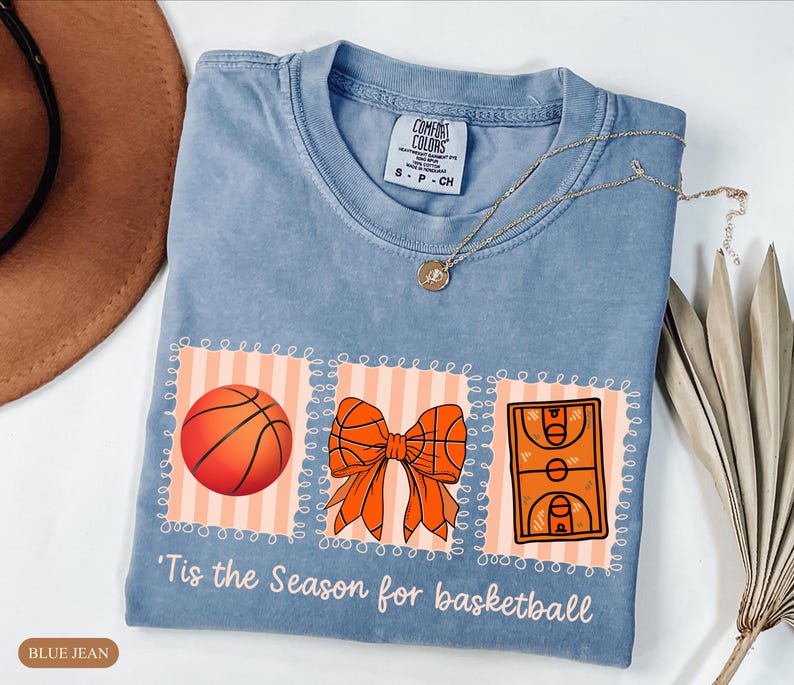 Peut inclure: T-shirt bleu jean clair avec des graphiques sur le th&egrave;me du basket-ball : un ballon, un n&oelig;ud et un terrain. Le texte "'Tis the Season for basketball" est imprim&eacute; en dessous. Un collier dor&eacute; est drap&eacute; sur le t-shirt.