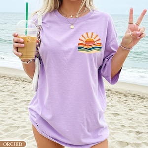 May include: Orchid purple t-shirt with a small graphic of a sunset over the ocean. The graphic features a yellow sun, orange and red rays, and blue, green, and orange waves. The shirt is a relaxed fit.