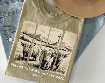 May include: A khaki t-shirt featuring a graphic print of three Highland cattle in a field with mountains and a cross in the background. The text reads, "FOLLOW THE WORD, NOT THE HERD" and "ISAIAH 8:11-3" in white lettering below the illustration.