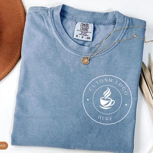 May include: A blue jean colored t-shirt with a crew neckline and a white circular graphic on the lower right side featuring a coffee cup icon and the text CUTOSM LOGO HERE. The shirt has a Comfort Colors brand label inside the collar.
