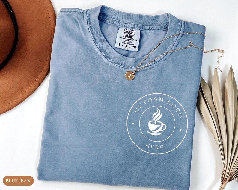 May include: A blue jean colored t-shirt with a crew neckline and a white circular graphic on the lower right side featuring a coffee cup icon and the text CUTOSM LOGO HERE. The shirt has a Comfort Colors brand label inside the collar.