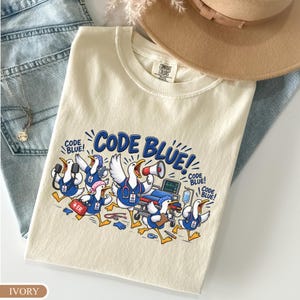 May include: An ivory t-shirt featuring a graphic of cartoon geese dressed as medical staff in blue scrubs, carrying medical equipment like a megaphone, stethoscope, and first aid kit, with the text "CODE BLUE!" printed in bold blue letters.