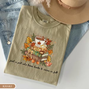 May include: A khaki t-shirt featuring a graphic of a stack of books, a cup of cocoa, autumn leaves, and a squirrel. The text reads, "just a girl who loves books & cocoa in fall." The shirt is displayed with a tan hat and blue denim fabric.
