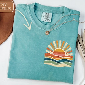 May include: A turquoise Comfort Colors t-shirt with a colourful sunset graphic. The graphic features a sun with orange and yellow rays, above layered orange, pink, and blue waves. A gold necklace with a pendant rests on the shirt.