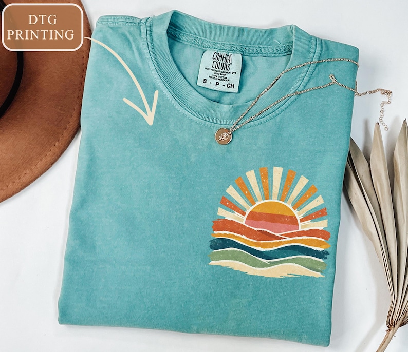 May include: A turquoise Comfort Colors t-shirt with a colourful sunset graphic. The graphic features a sun with orange and yellow rays, above layered orange, pink, and blue waves. A gold necklace with a pendant rests on the shirt.