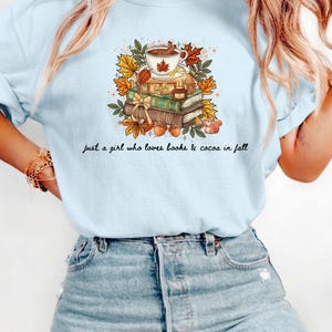 May include: A light blue t-shirt featuring a graphic of a stack of books topped with a mug of cocoa, autumn leaves, acorns, and a small squirrel. The text below the illustration reads, "just a girl who loves books & cocoa in fall."