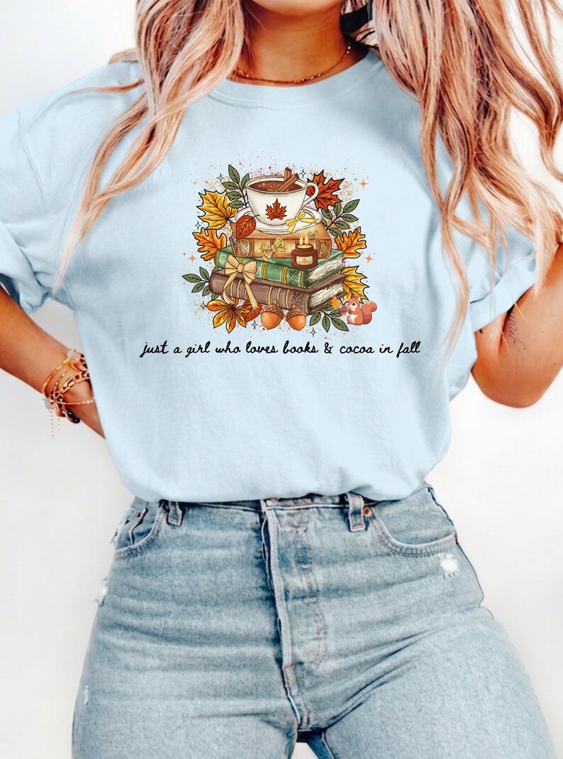 May include: A light blue t-shirt featuring a graphic of a stack of books topped with a mug of cocoa, autumn leaves, acorns, and a small squirrel. The text below the illustration reads, "just a girl who loves books & cocoa in fall."
