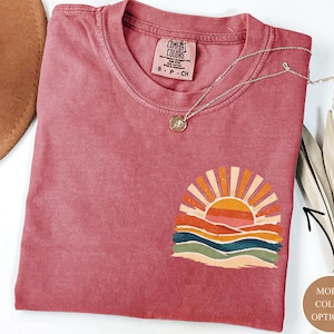 May include: A dusky pink Comfort Colors t-shirt with a sunset graphic. The graphic shows an orange sun with rays over colourful waves. A silver necklace with a pendant is on the shirt.