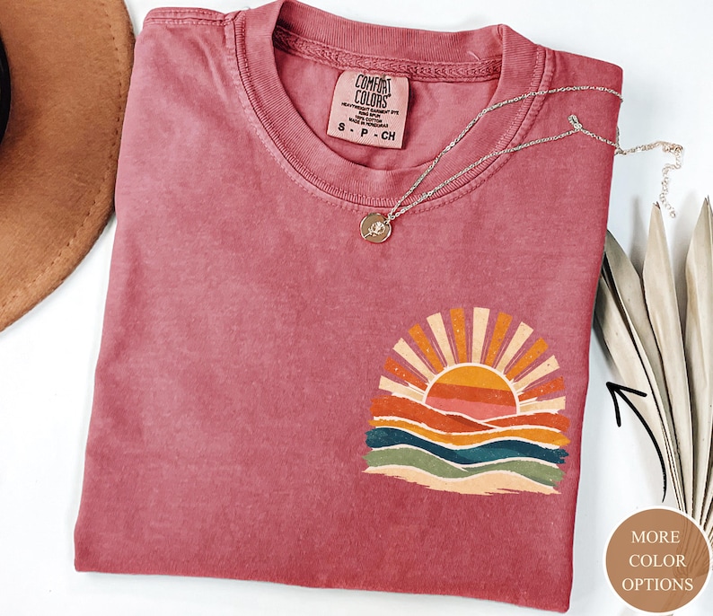 May include: A dusky pink Comfort Colors t-shirt with a sunset graphic. The graphic shows an orange sun with rays over colourful waves. A silver necklace with a pendant is on the shirt.