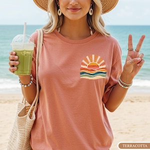 May include: Terracotta-coloured t-shirt featuring a sunset graphic. The graphic shows a sun with orange and yellow rays above blue and green waves. The shirt is worn on a beach with a straw hat and a tote bag. The word "TERRACOTTA" is at the bottom.