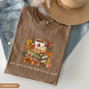 May include: A brown t-shirt featuring a graphic of a stack of books, a cup of cocoa, and autumn leaves. The text reads, "just a girl who loves books & cocoa in fall." The shirt is layered over light blue denim fabric and a tan wide-brimmed hat.