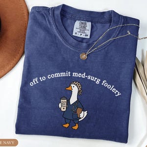 May include: A navy blue t-shirt featuring a graphic of a white duck wearing a leopard print scrub cap, a stethoscope, and blue scrubs. The duck holds a cup labeled Charts Meds Chaos. The text reads off to commit med-surg foolery in white.