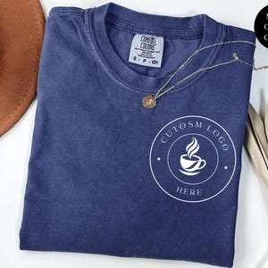 May include: A navy blue short-sleeve t-shirt with a crew neckline and a white circular graphic on the chest featuring a coffee cup icon and the text "CUTOSM LOGO HERE". The shirt has a Comfort Colors brand label at the inner neck showing size S.