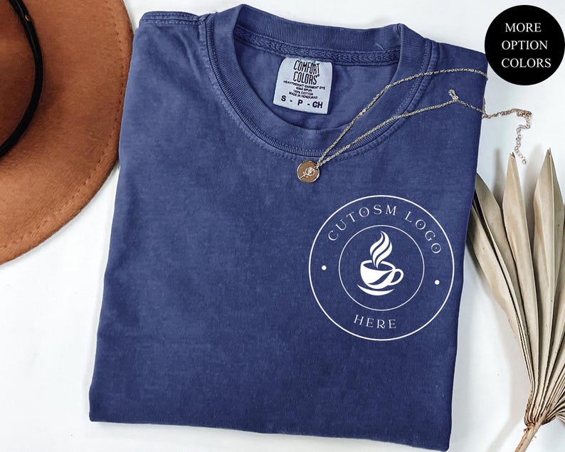 May include: A navy blue short-sleeve t-shirt with a crew neckline and a white circular graphic on the chest featuring a coffee cup icon and the text "CUTOSM LOGO HERE". The shirt has a Comfort Colors brand label at the inner neck showing size S.
